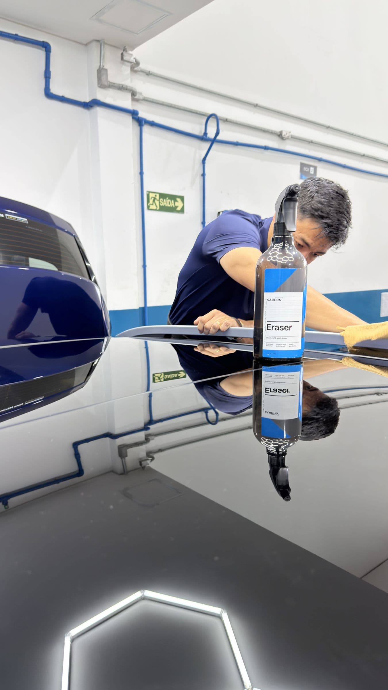 Profissional da equipe aplicando produto CARPRO sobre o teto de um carro, com o próprio reflexo na pintura espelhada.