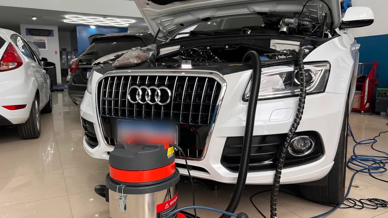 Audi branco com o capô aberto na oficina, com o extrator conectado ao compartimento do motor durante o diagnóstico do sistema de combustível.