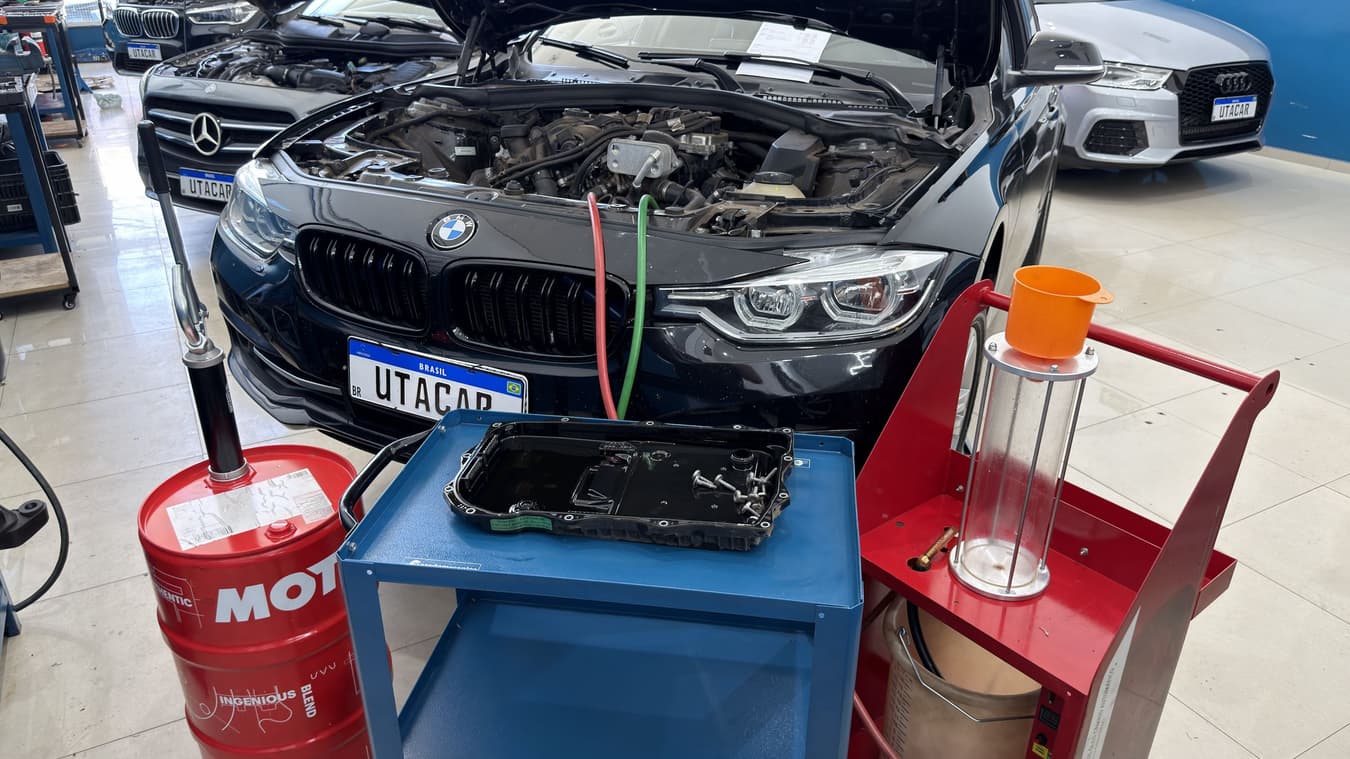 BMW com o capô aberto na Utacar, mangueiras da máquina de troca de fluido conectadas e o cárter do câmbio sobre o carrinho ao lado.