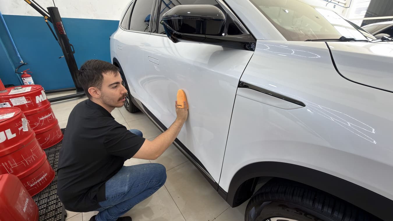 Técnico agachado aplica o revestimento na lateral do SUV branco, com os barris de produto ao fundo no box da Utacar.
