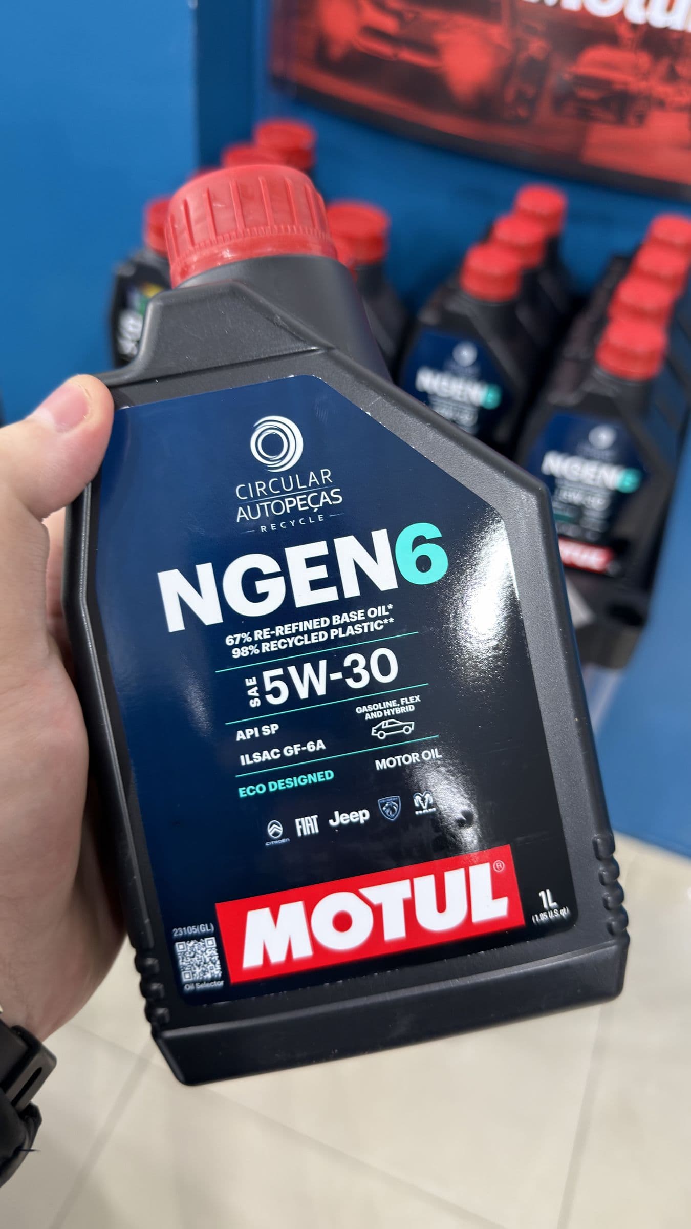 Mão segura o frasco de 1 litro do Motul NGEN 6 5W-30 Circular Autopeças, com o rótulo frontal indicando API SP, ILSAC GF-6A e 67% de óleo básico rerrefinado.