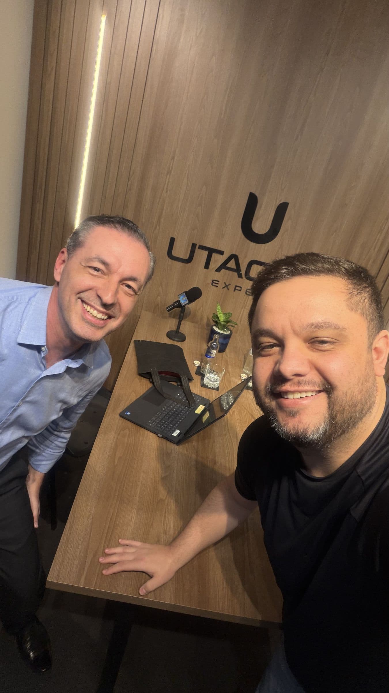 Gustavo Agiessi e o advogado Mauricio Plinta lado a lado na mesa do UtaPapo, com o painel de madeira e o logotipo da Utacar ao fundo, microfone, notebook e copos sobre a mesa.