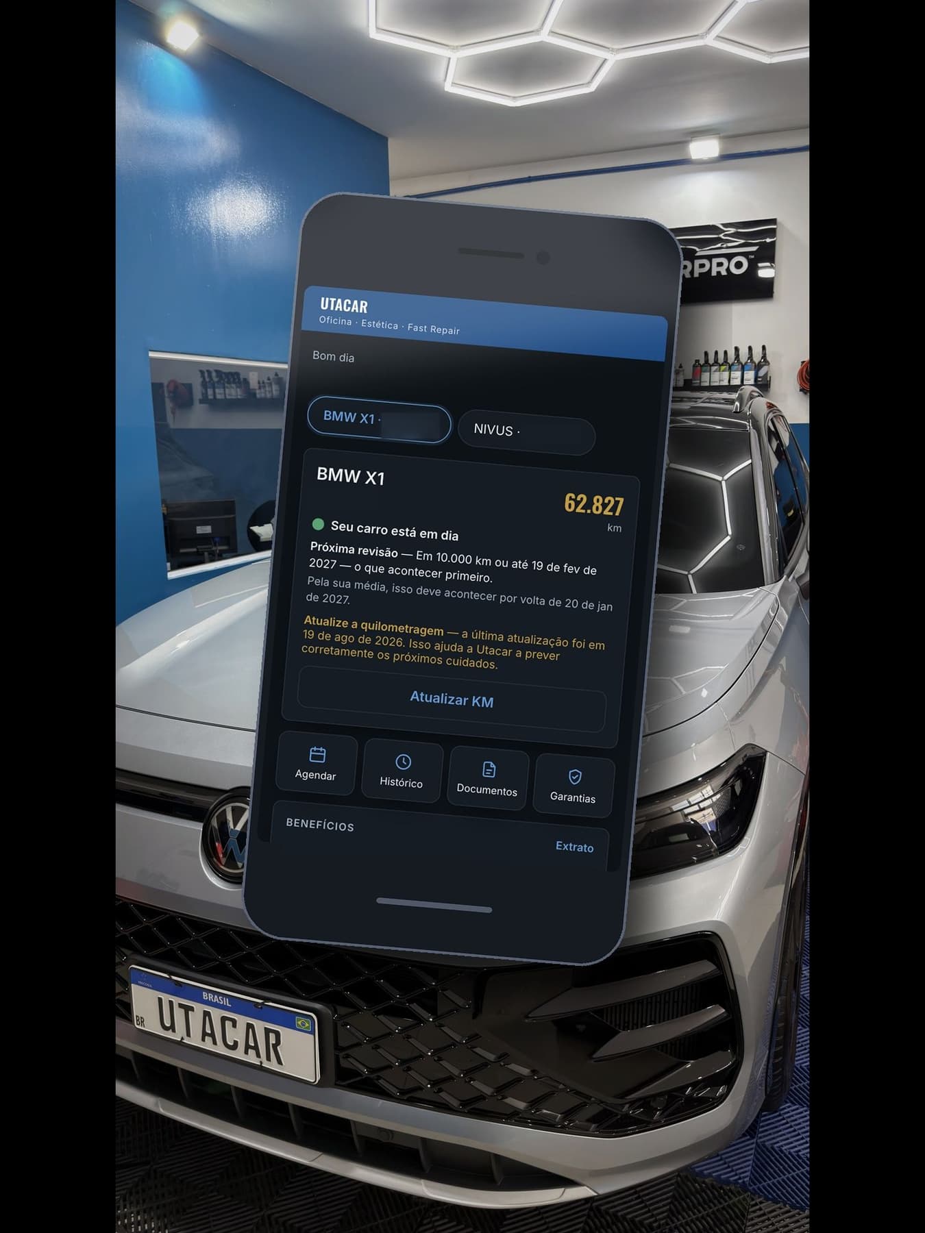 Tela inicial do aplicativo da Utacar num celular, sobre a foto de um carro na oficina: os veículos do cliente, a quilometragem de 62.827 km, o aviso de que o carro está em dia, a previsão da próxima revisão e os atalhos Agendar, Histórico, Documentos e Garantias. As placas aparecem ocultadas.