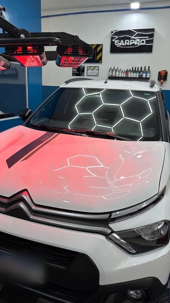 Citroën branco no box de estética sob as lâmpadas de infravermelho, com a iluminação hexagonal refletida no capô protegido e o painel da CARPRO na parede ao fundo.