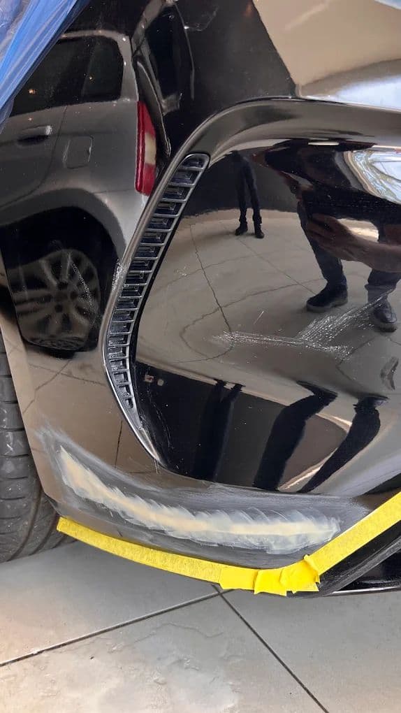 Canto de para-choque preto com o dano lixado e fita de proteção amarela, preparado para pintura localizada de Fast Repair.