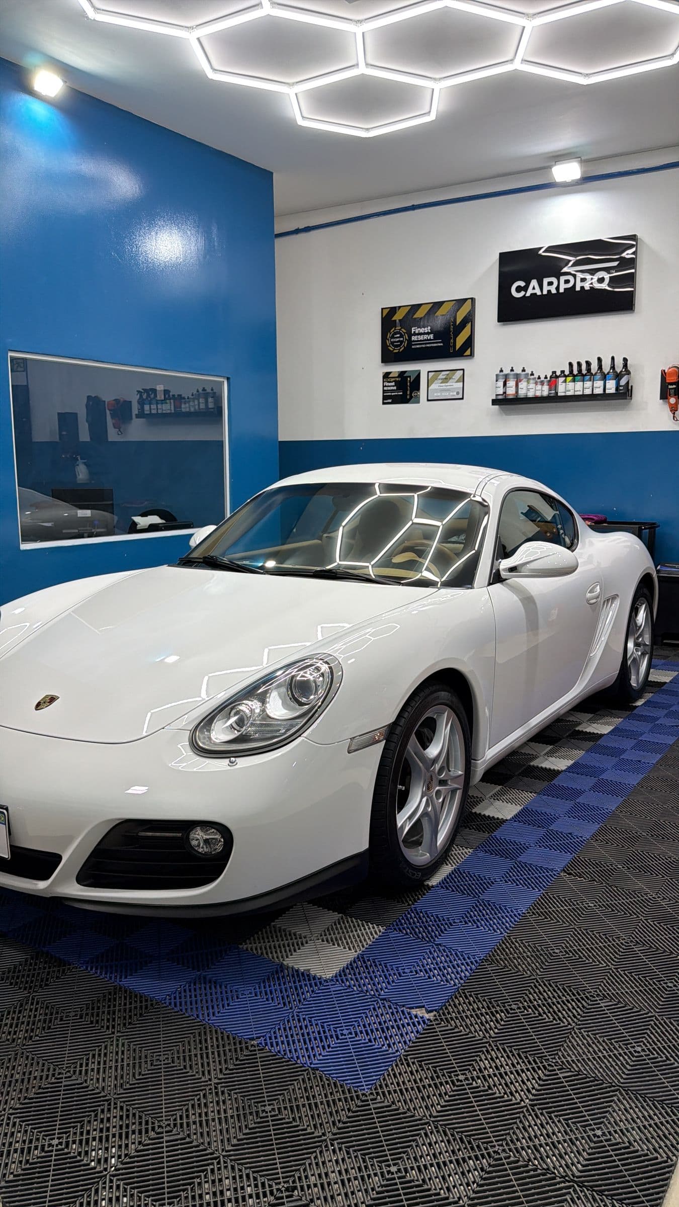Porsche Cayman branco no box exclusivo de estética, visto de três quartos sob a iluminação hexagonal, com o painel da CARPRO e os certificados na parede.