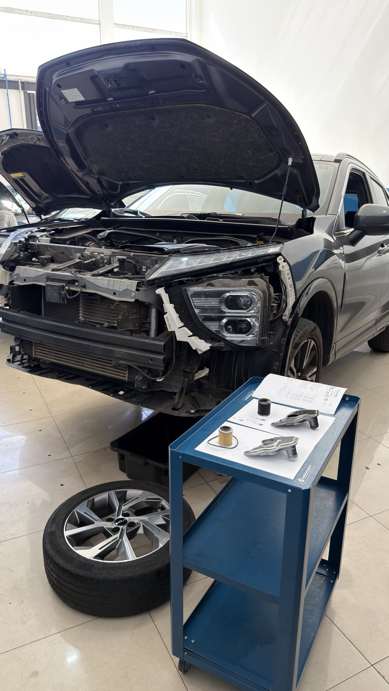 Carro com o para-choque dianteiro removido para pintura, ao lado do carrinho com as peças da transmissão.