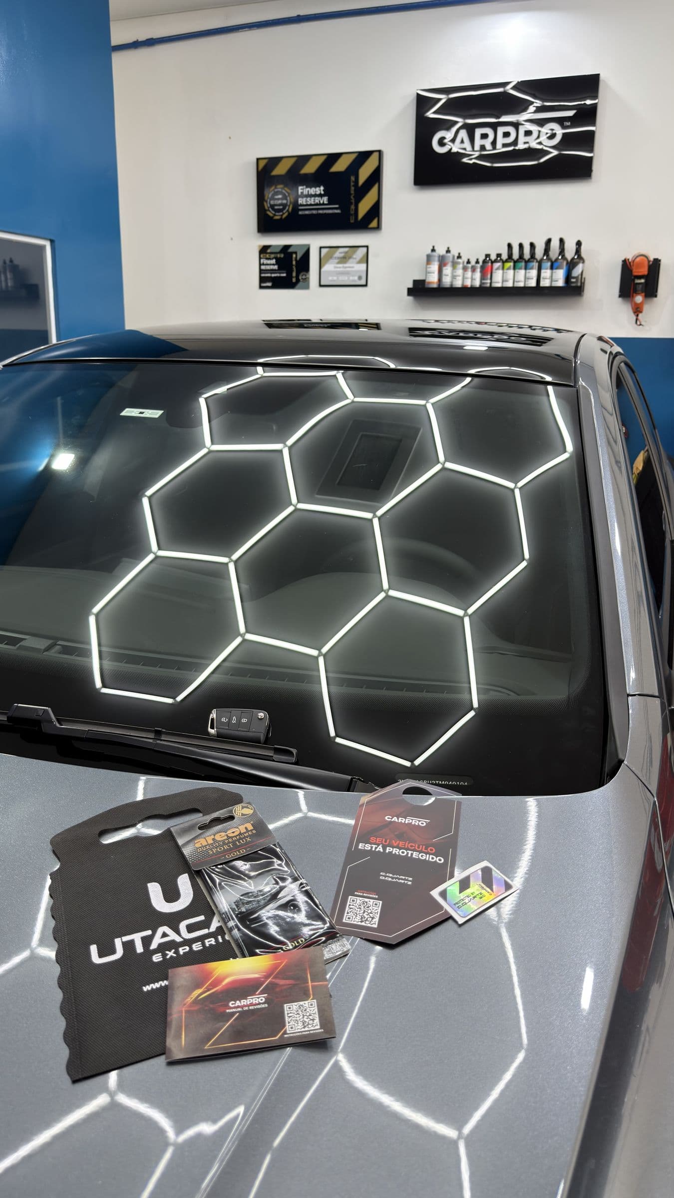Para-brisa de um carro protegido refletindo a iluminação hexagonal, com o rodo UTACAR e os cartões de cuidado da CARPRO apoiados no capô.
