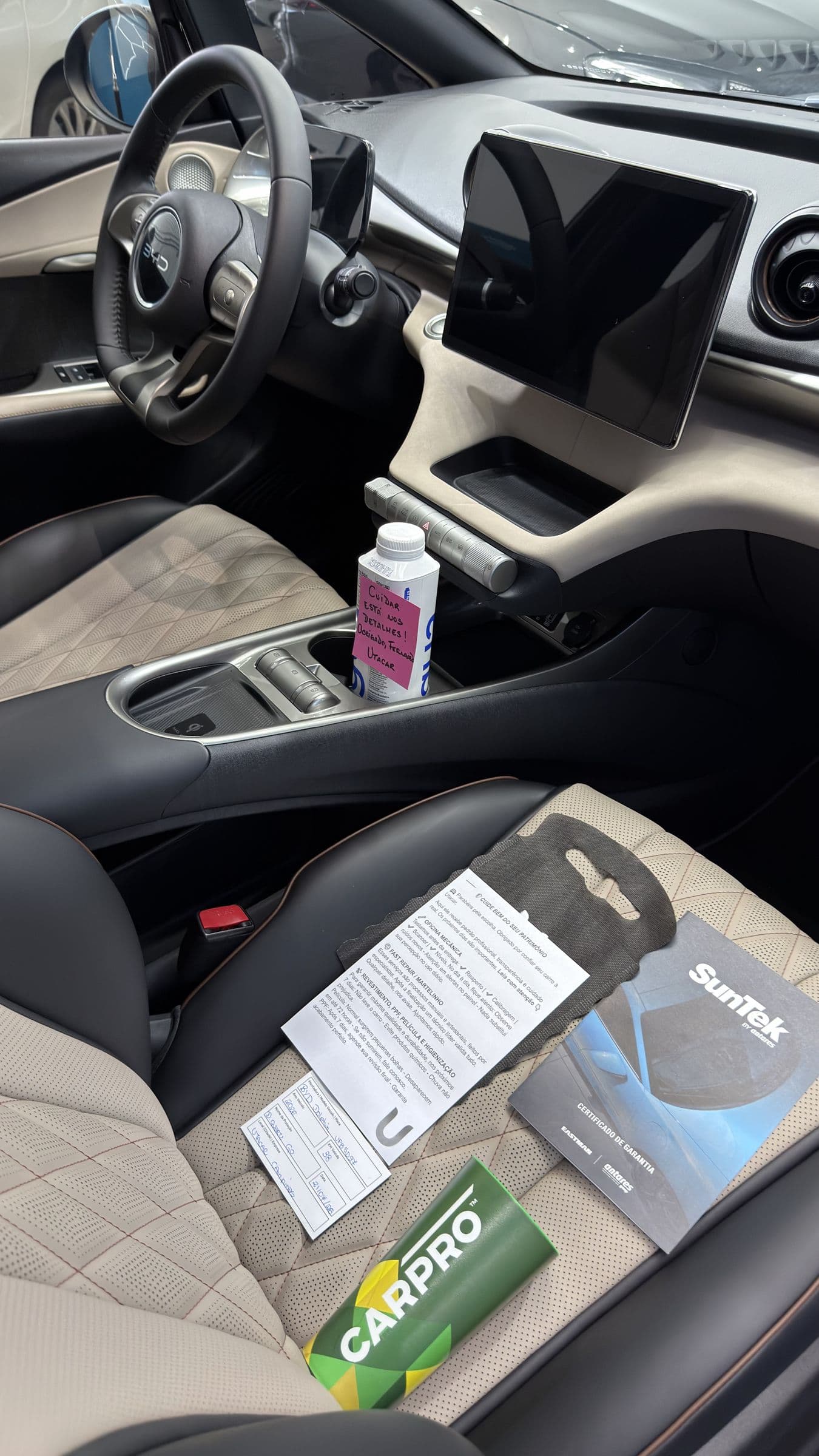 Interior claro de um BYD com o kit de entrega da Utacar, o certificado SunTek e um bilhete escrito à mão sobre o banco.