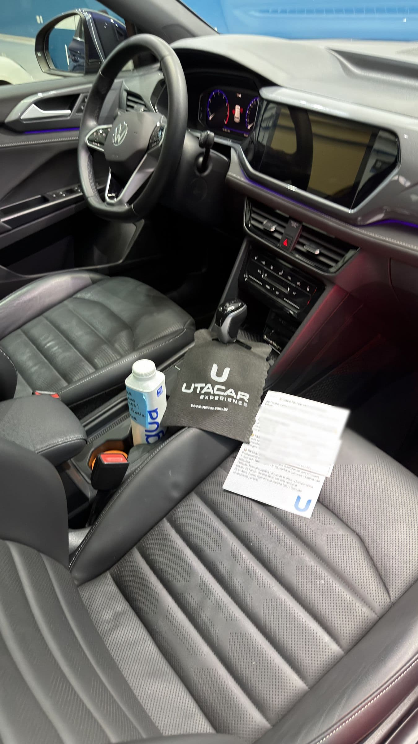 Interior de um Volkswagen T-Cross pronto para a entrega, com a flanela UTACAR sobre o câmbio, uma garrafa de água de cortesia e o cartão de orientações no banco.