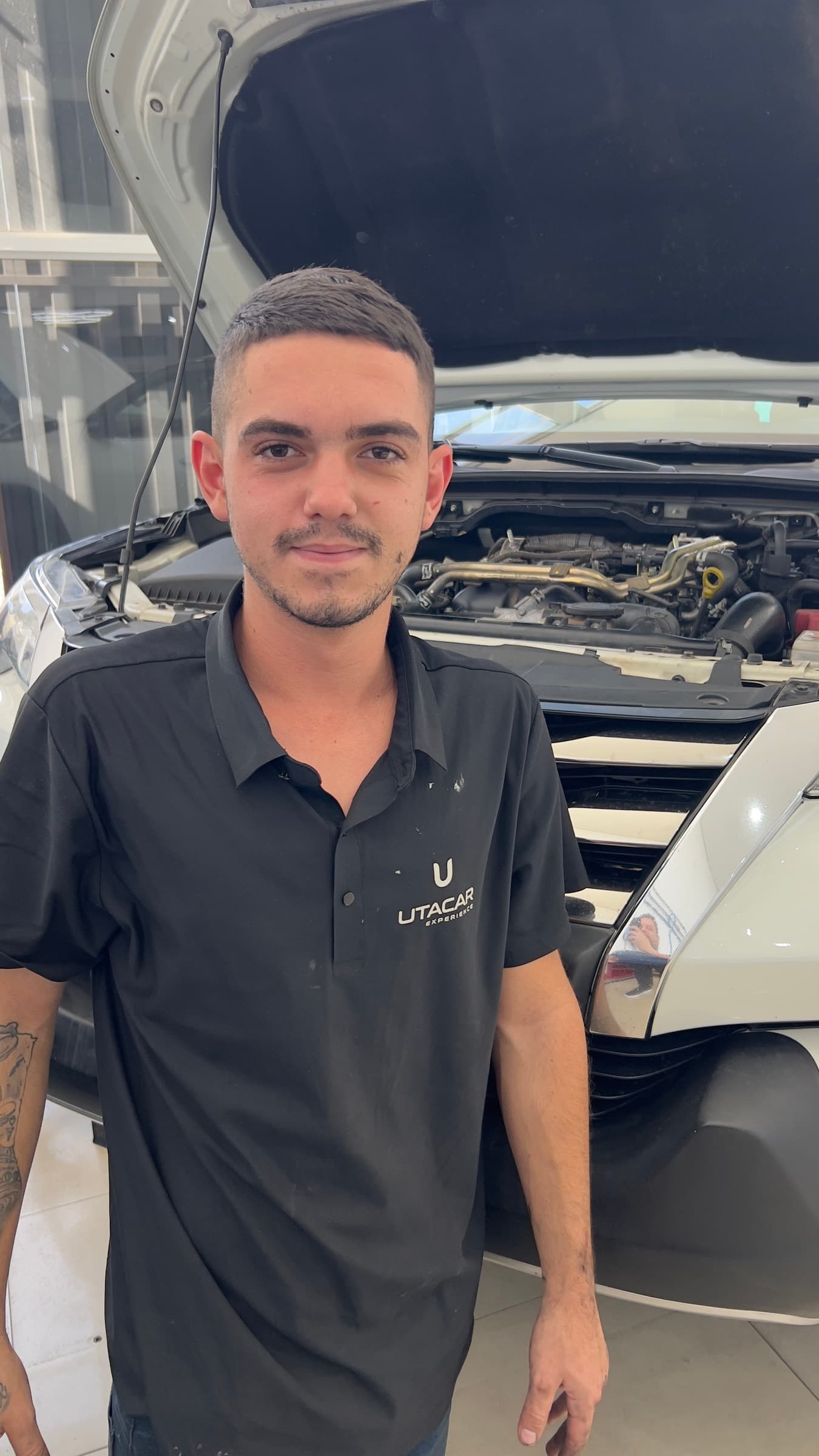 Davi Lopes, técnico de oficina da Utacar, de uniforme, diante de um carro com o capô aberto na oficina.