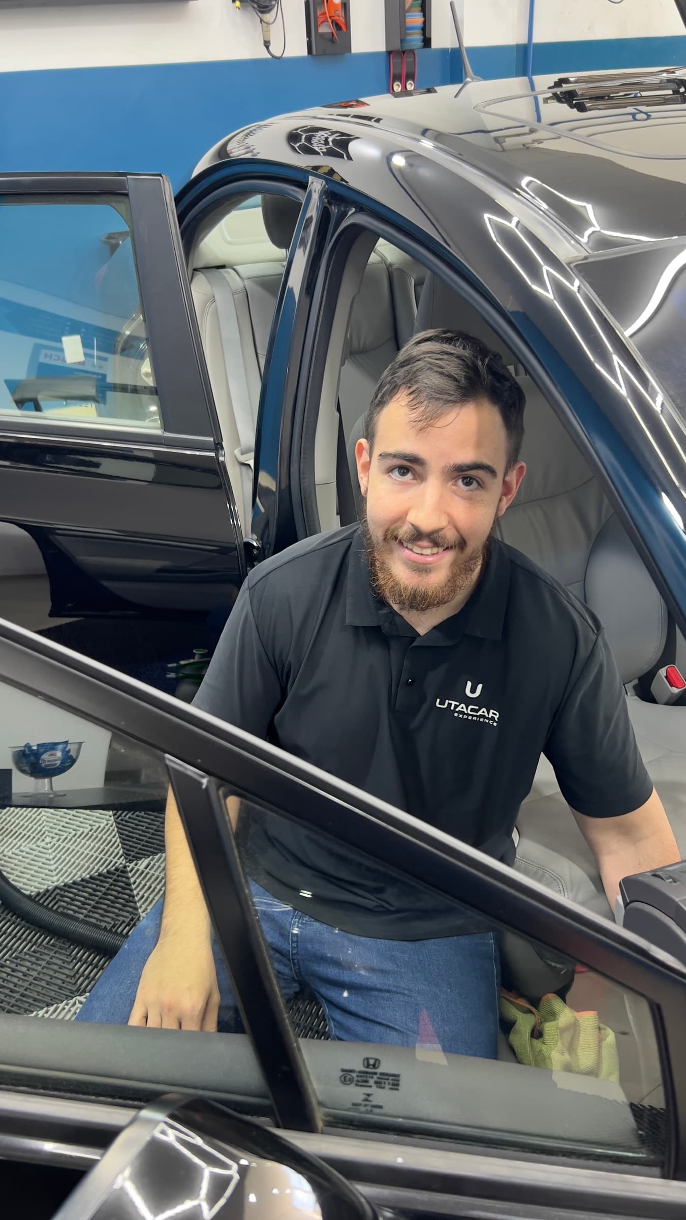 Gabriel Munhoz, técnico em estética automotiva da Utacar, de uniforme, ao lado da porta aberta de um carro no box.