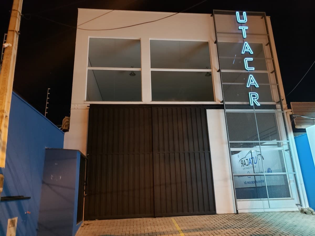 Fachada da Utacar à noite, com o letreiro vertical de neon aceso sobre o portão fechado.