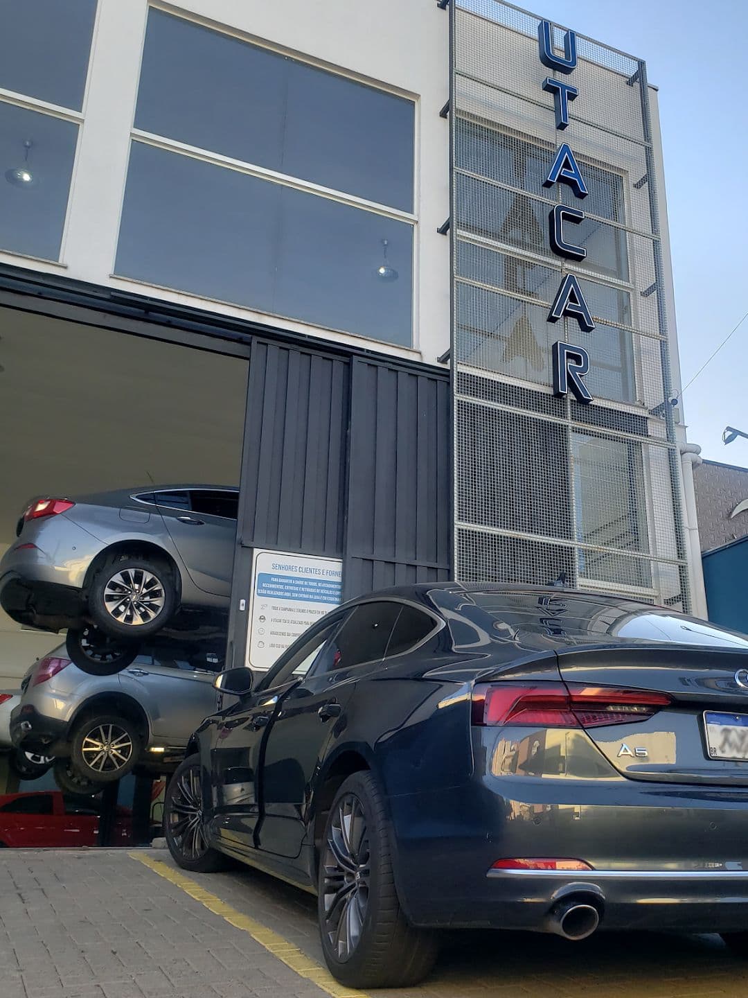 Fachada da Utacar vista da calçada, com o letreiro vertical no alto, um carro sobre o elevador dentro da oficina e um Audi estacionado na frente.