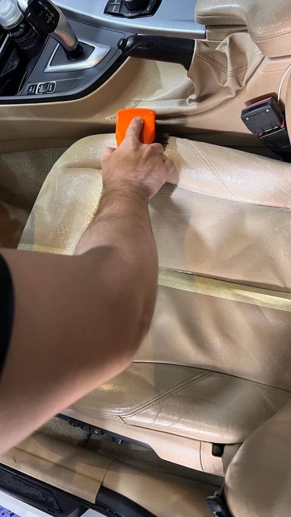 Mão da equipe aplicando produto de proteção sobre o banco de couro claro com um aplicador laranja.
