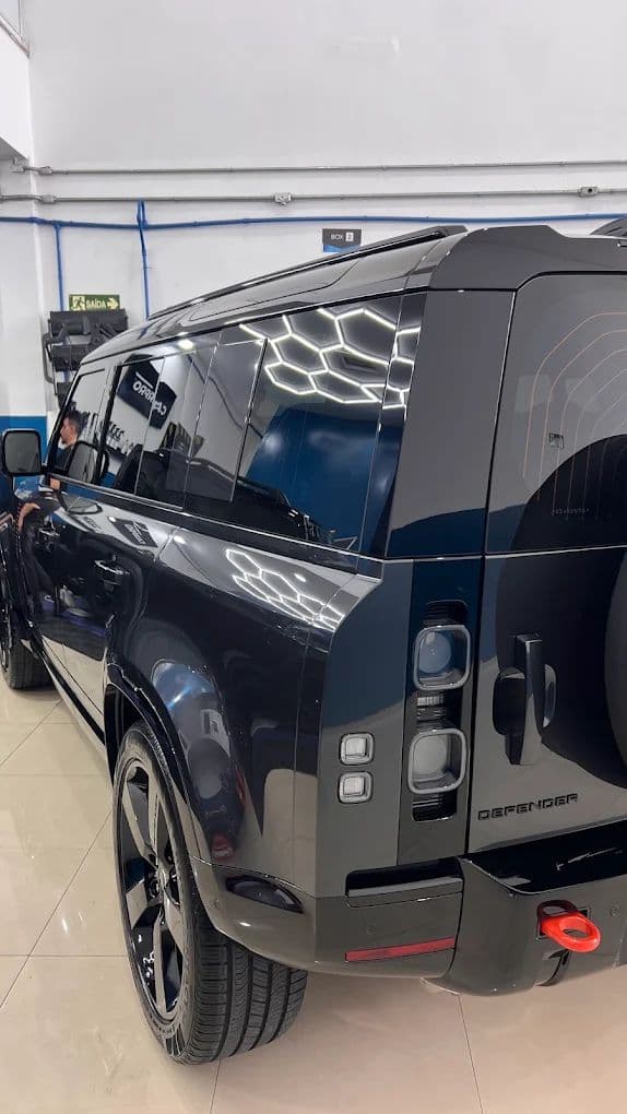 Land Rover Defender preto visto de trás no box de estética, com a iluminação hexagonal do teto refletida na lataria.
