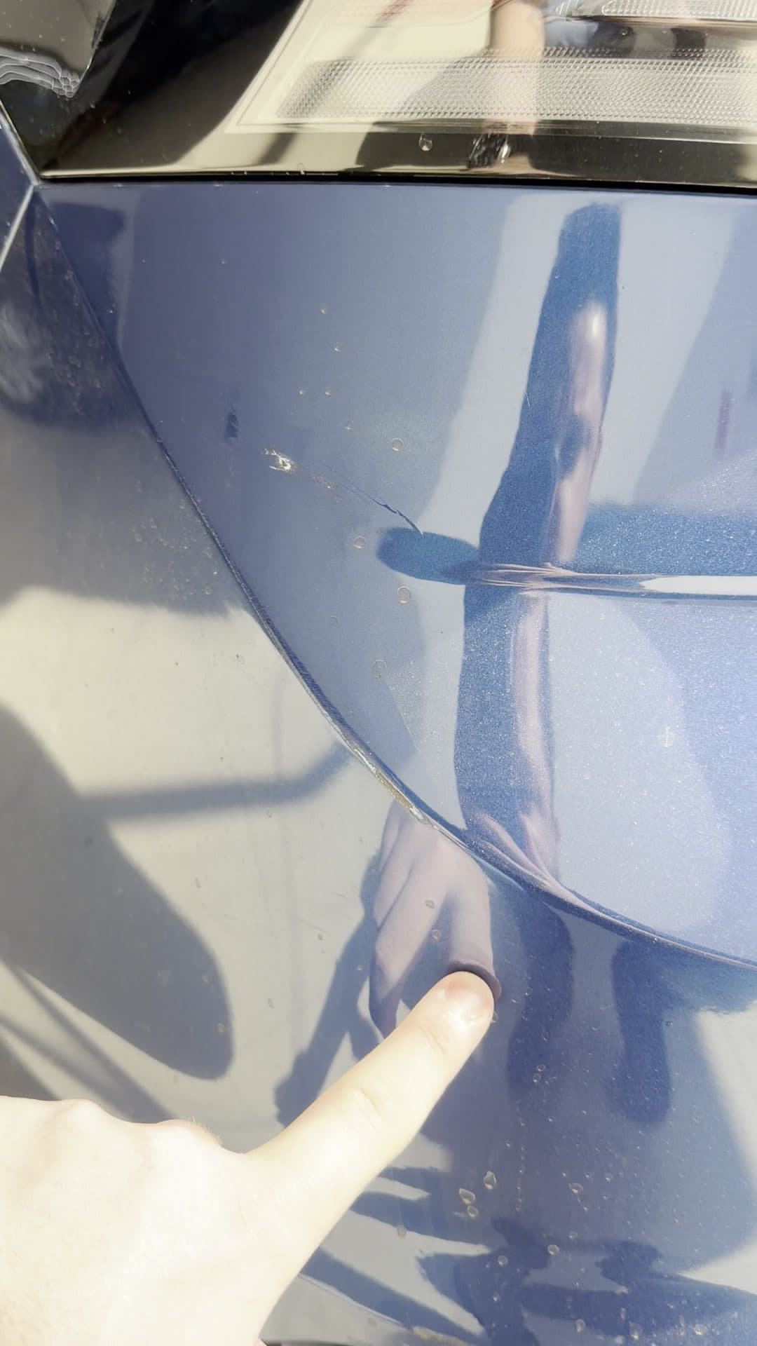 Dedo apontando o amassado na lateral traseira de um carro, antes do reparo.