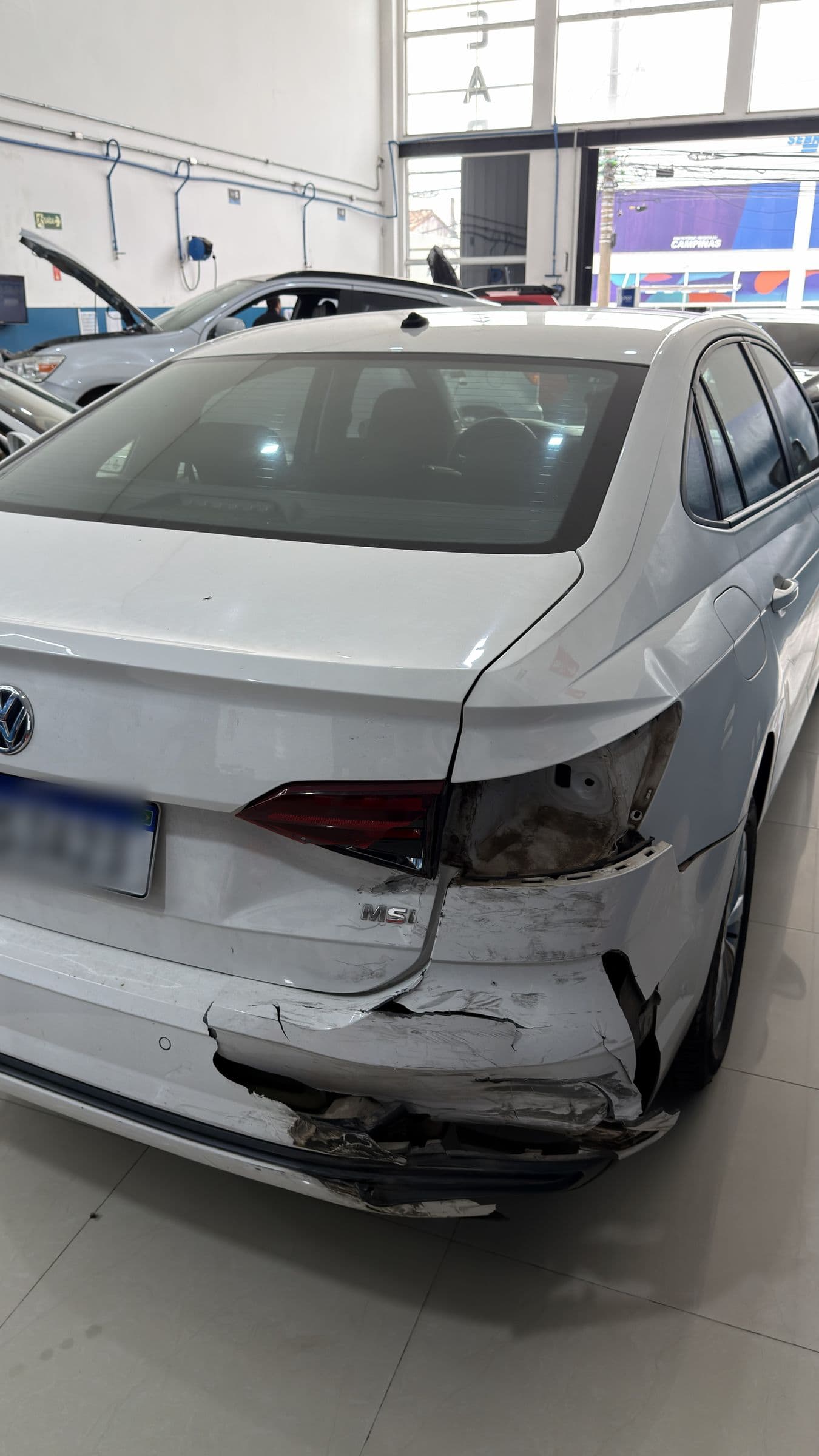 Traseira de um Volkswagen branco com o para-choque e a lateral amassados depois de uma colisão, dentro da oficina.
