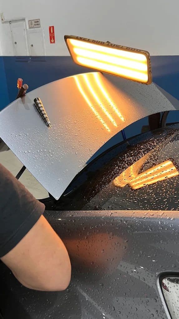 Barra de luz de LED refletindo linhas sobre o capô cinza de um carro durante o martelinho de ouro, com a ferramenta de repuxo apoiada na peça.