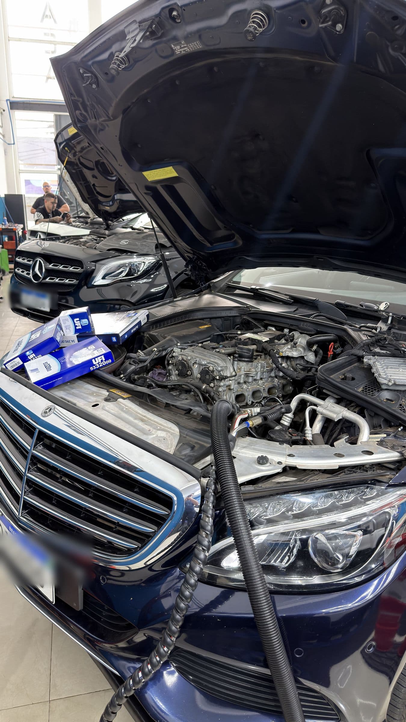 Compartimento do motor de uma Mercedes-Benz aberto para revisão, com as caixas dos filtros novos apoiadas no para-lama e outra Mercedes ao fundo.