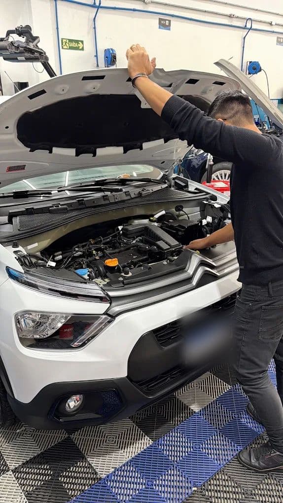 Profissional da equipe levantando o capô de um Citroën branco na oficina, com o compartimento do motor à mostra e a sinalização do box ao fundo.