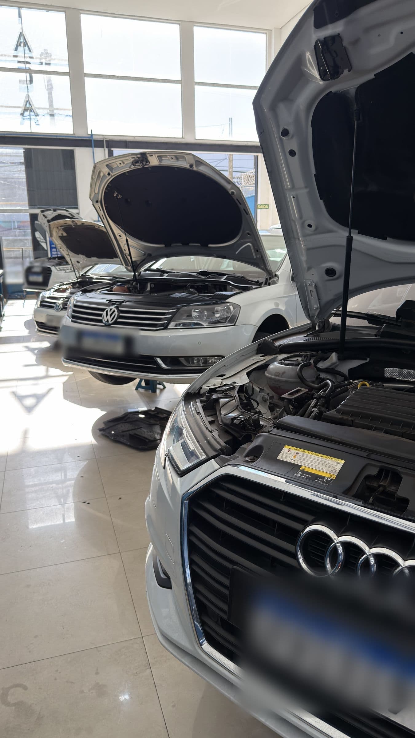 Três carros com o capô aberto no salão da oficina: um Audi em primeiro plano, um Volkswagen Passat sobre o elevador ao centro e um terceiro ao fundo, com a claraboia iluminando o piso claro.