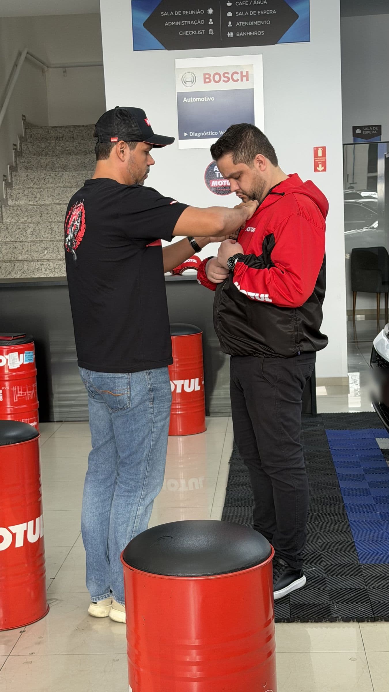 Entrega do agasalho Motul no balcão da Utacar, com o painel da Bosch e os tambores de lubrificante ao fundo.