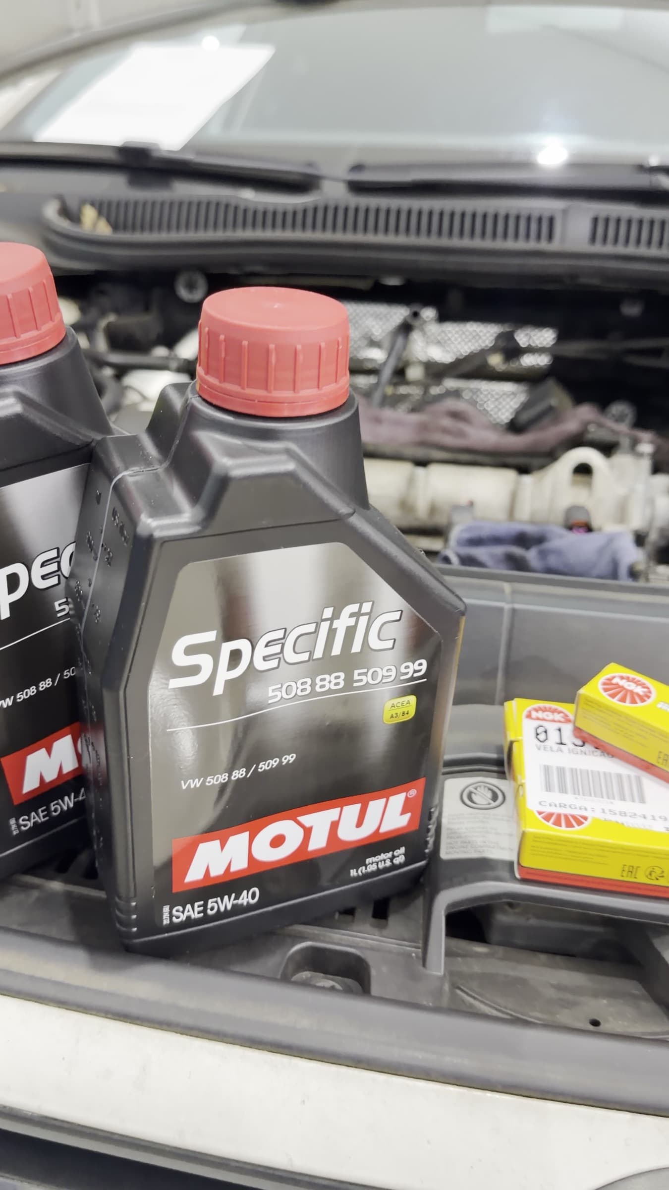 Frascos de óleo Motul Specific 5W-40, na especificação VW 508 88 / 509 99, apoiados no compartimento do motor ao lado das caixas de velas NGK.