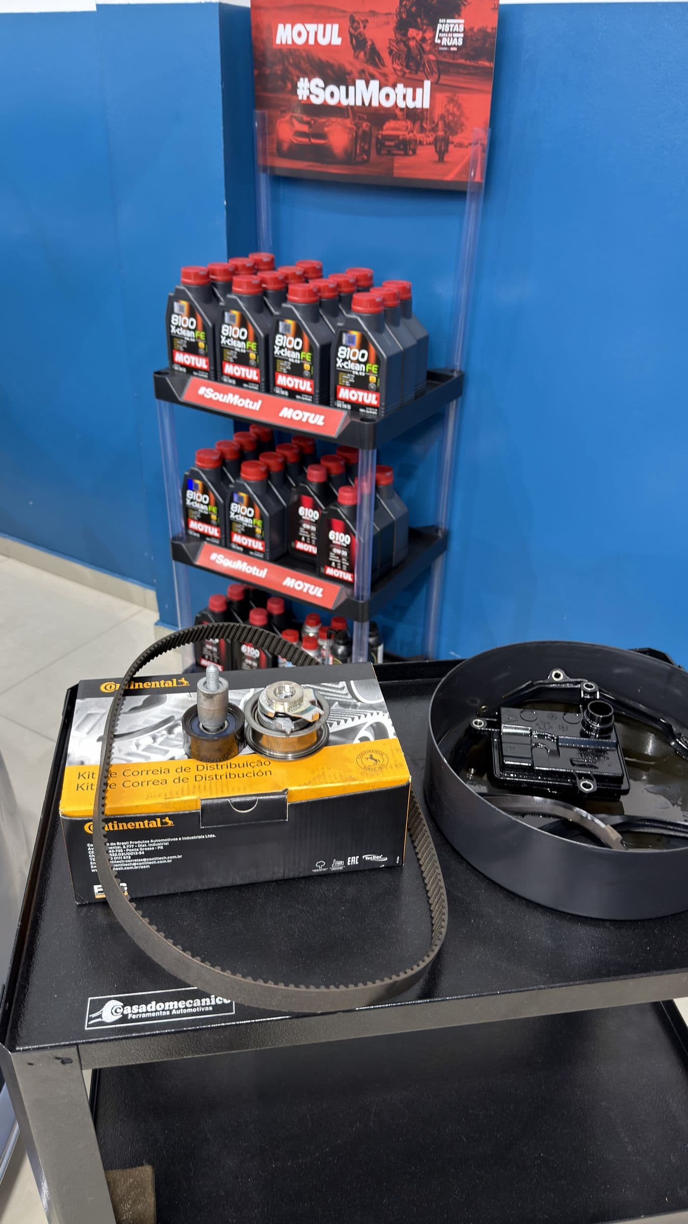 Kit de correia de distribuição Continental na caixa aberta sobre o carrinho de ferramentas, ao lado do cárter e do filtro, com a prateleira de lubrificantes Motul ao fundo.
