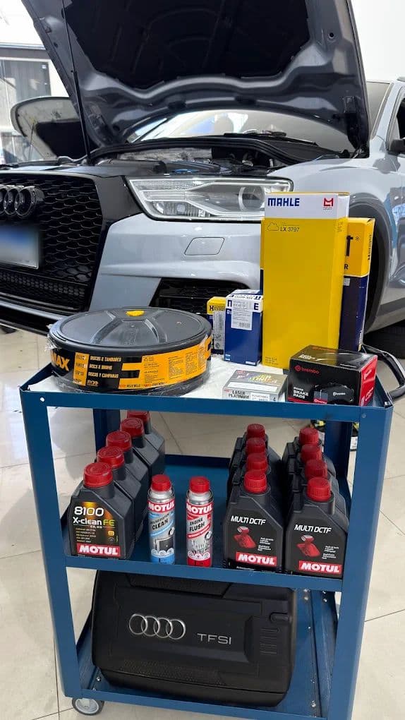 Carrinho com os frascos de lubrificante Motul, filtros e demais itens separados para a revisão, com um Audi de capô aberto ao fundo.