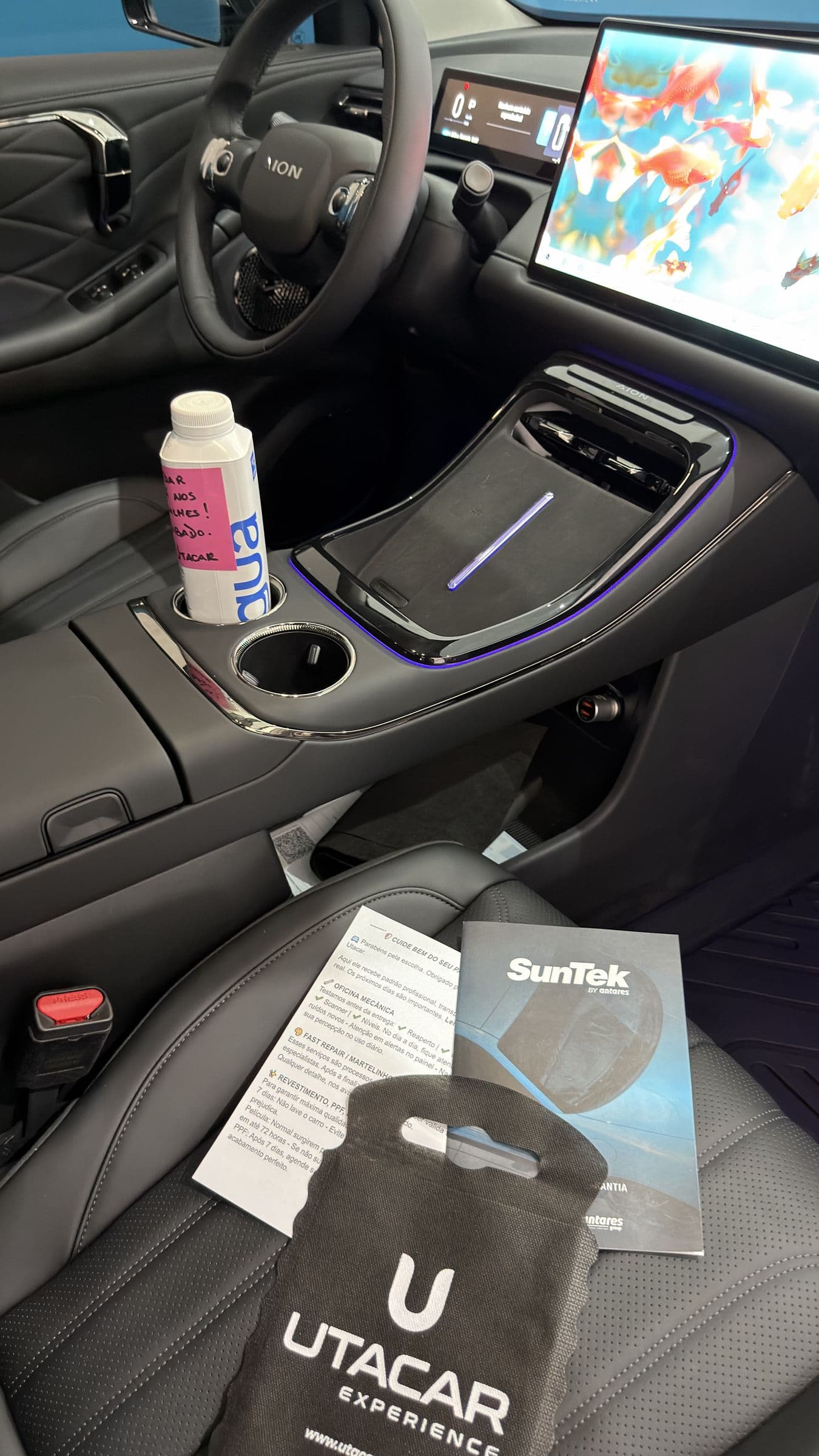 Interior de um SUV elétrico pronto para a entrega, com a flanela UTACAR no banco, o certificado de garantia SunTek e uma garrafa de água de cortesia com bilhete escrito à mão.