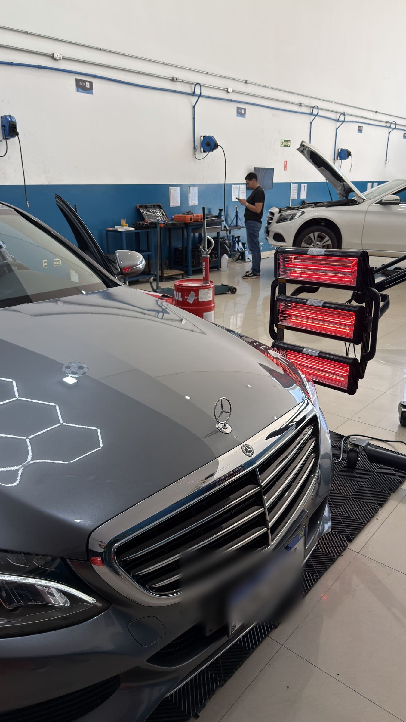 Mercedes cinza estacionado ao lado da torre de lâmpadas infravermelhas usada para curar a pintura.