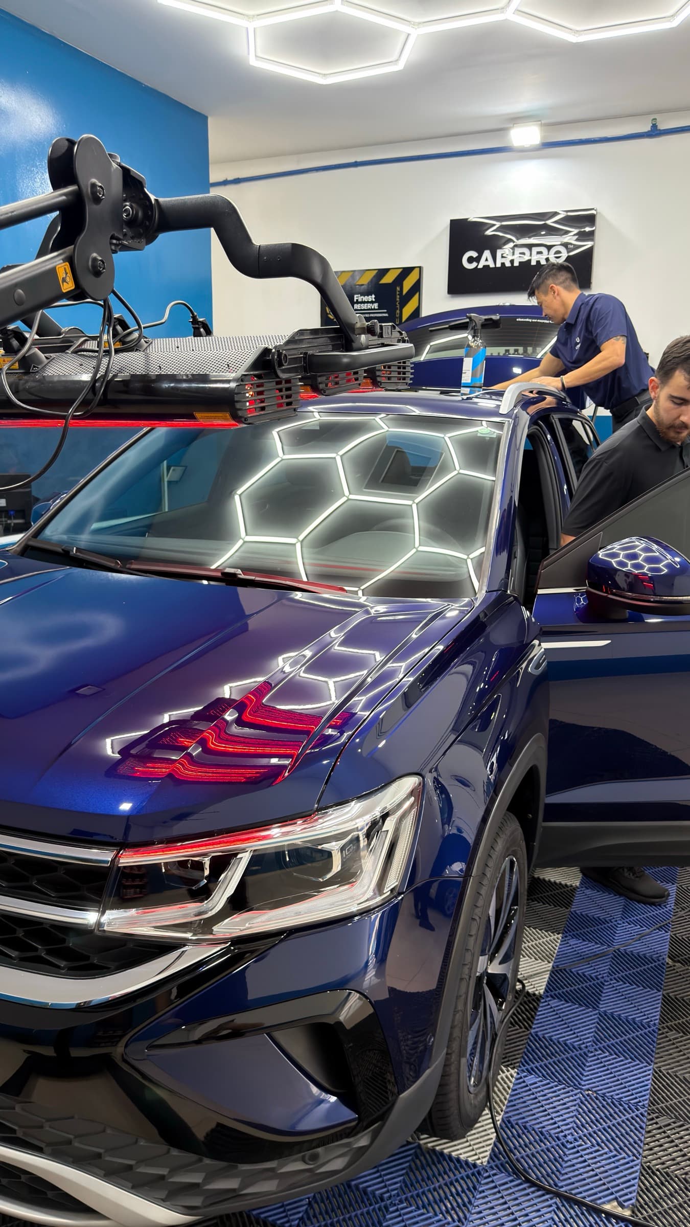 Dois profissionais da equipe trabalhando na pintura de um SUV azul sob o painel de luzes hexagonais do box de estética.