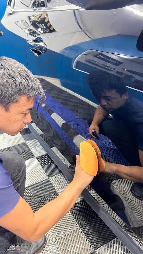Dois profissionais da equipe polindo a lateral de um carro azul, um com a politriz na mão e o outro conferindo o resultado de perto.