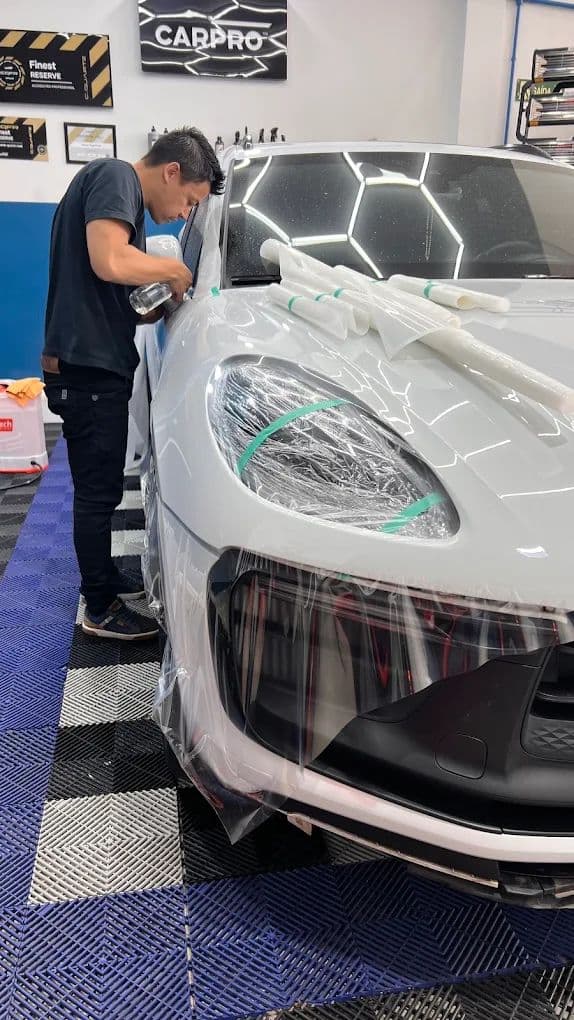 Profissional da equipe aplicando a película de PPF sobre o para-choque dianteiro de um Porsche branco no box de estética.
