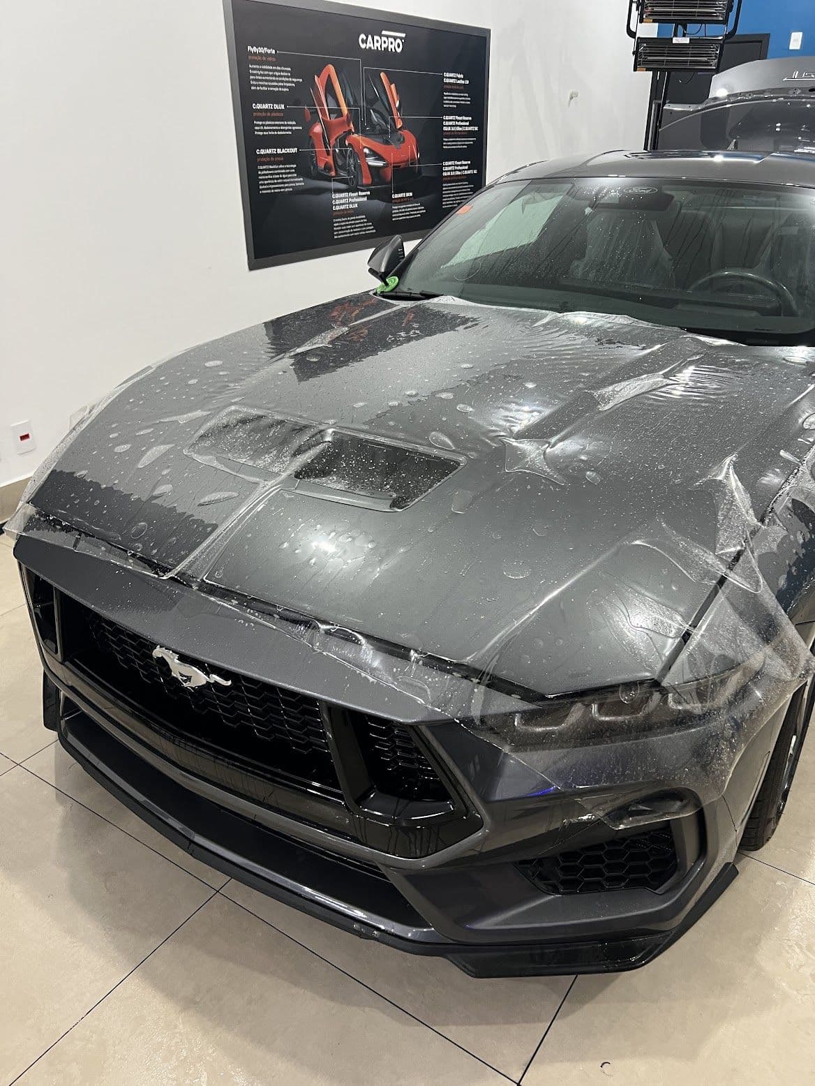 Película de PPF sendo assentada sobre o capô de um Ford Mustang cinza no box de estética, com o painel da CARPRO na parede ao fundo.