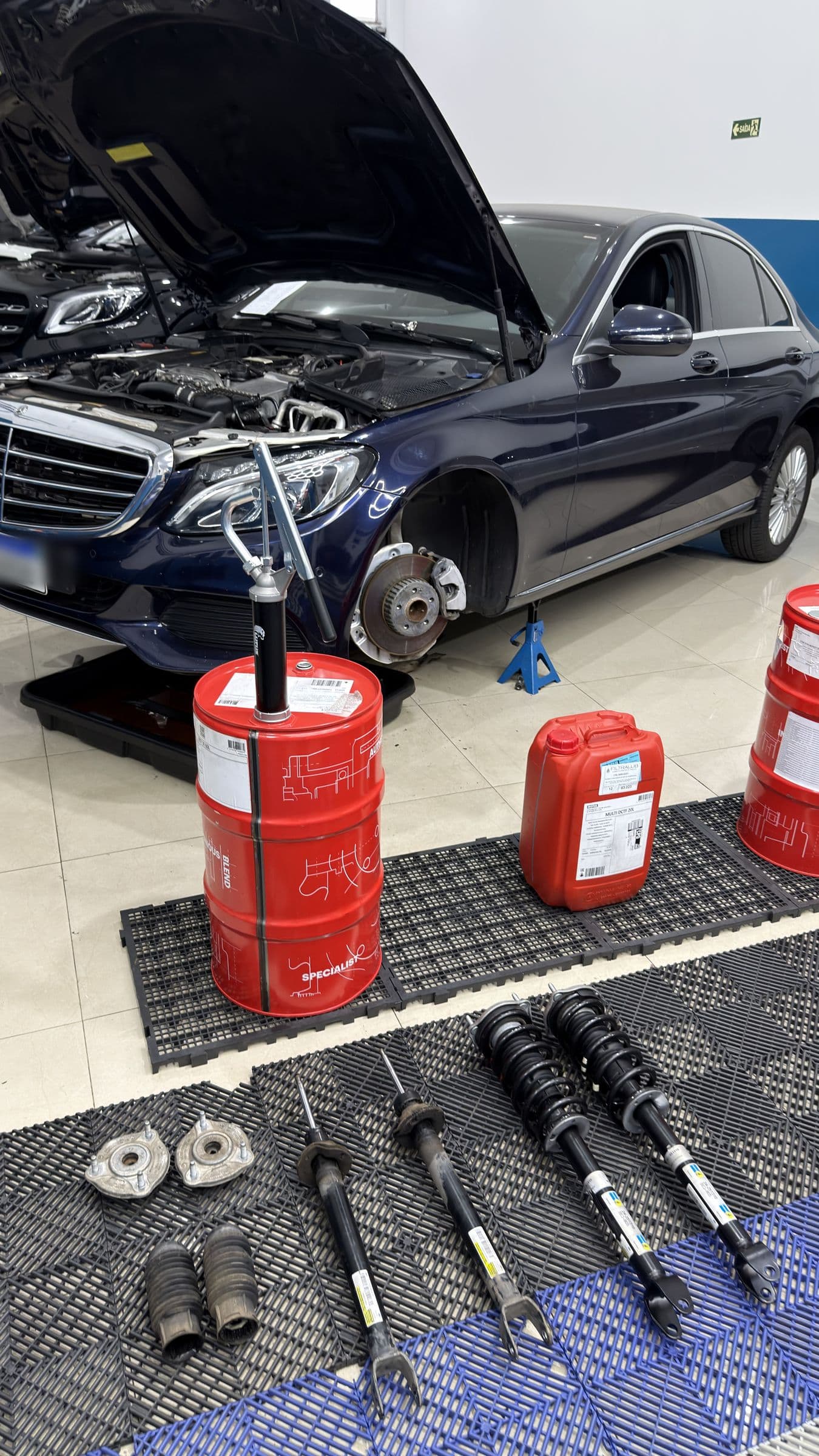 Mercedes-Benz sobre o cavalete com a roda removida, e os amortecedores Bilstein novos alinhados no piso ao lado dos tambores de lubrificante.