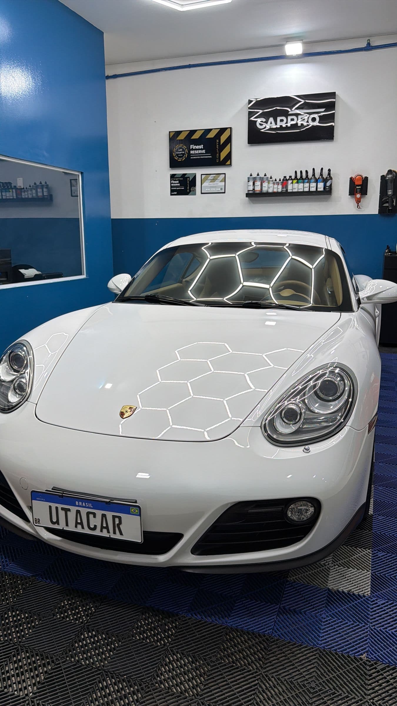 Porsche Cayman branco visto de frente no box de estética, sob a iluminação hexagonal, com a placa de cobertura da UTACAR.