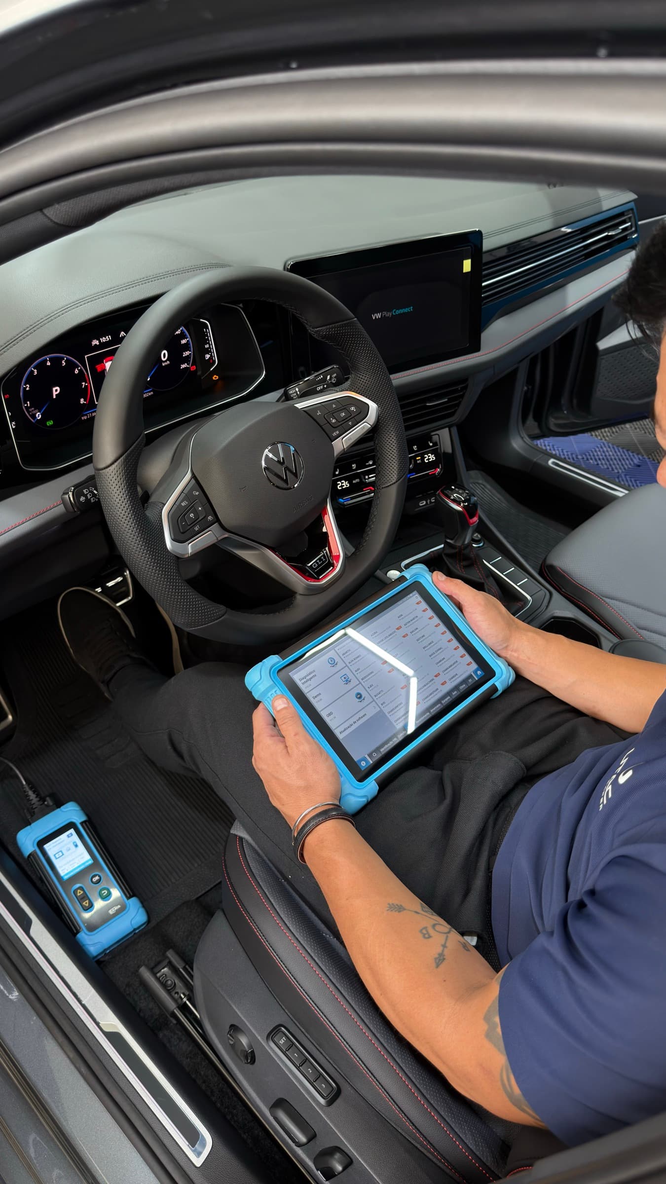 Técnico sentado ao volante de um Volkswagen com o tablet de diagnóstico nas mãos, o módulo de interface ligado no soquete e o painel do carro aceso.