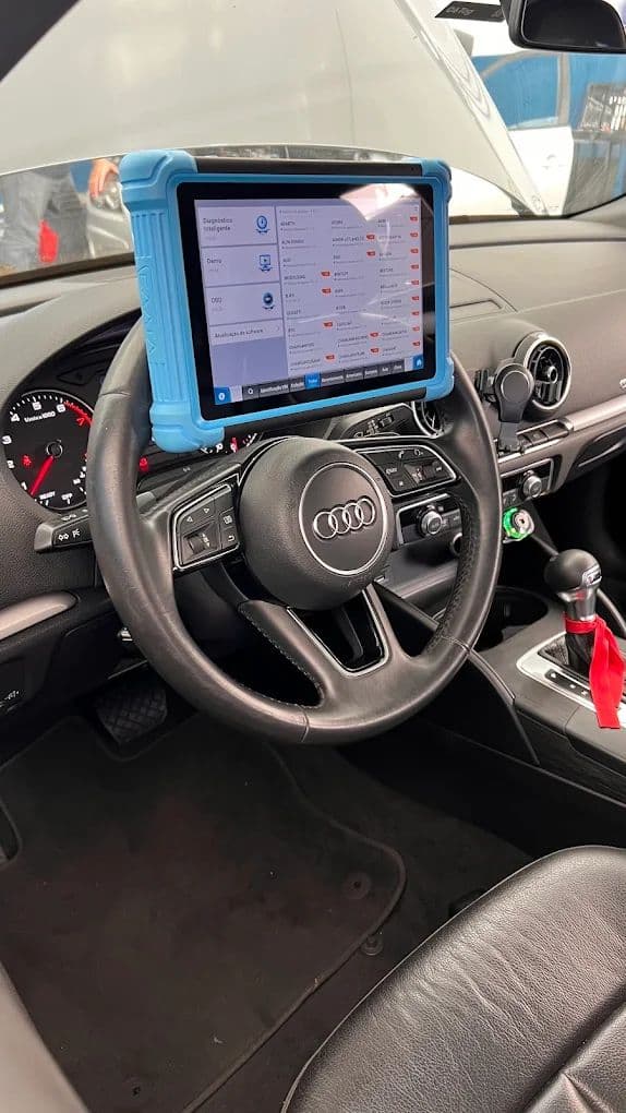 Tablet de diagnóstico preso ao volante de um Audi, com a tela de leitura dos módulos aberta e o painel do carro ao fundo.