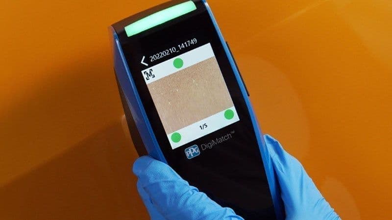 Espectrofotômetro PPG DigiMatch segurado por uma mão com luva azul sobre a pintura laranja de um carro, com a leitura da cor aberta na tela do aparelho.