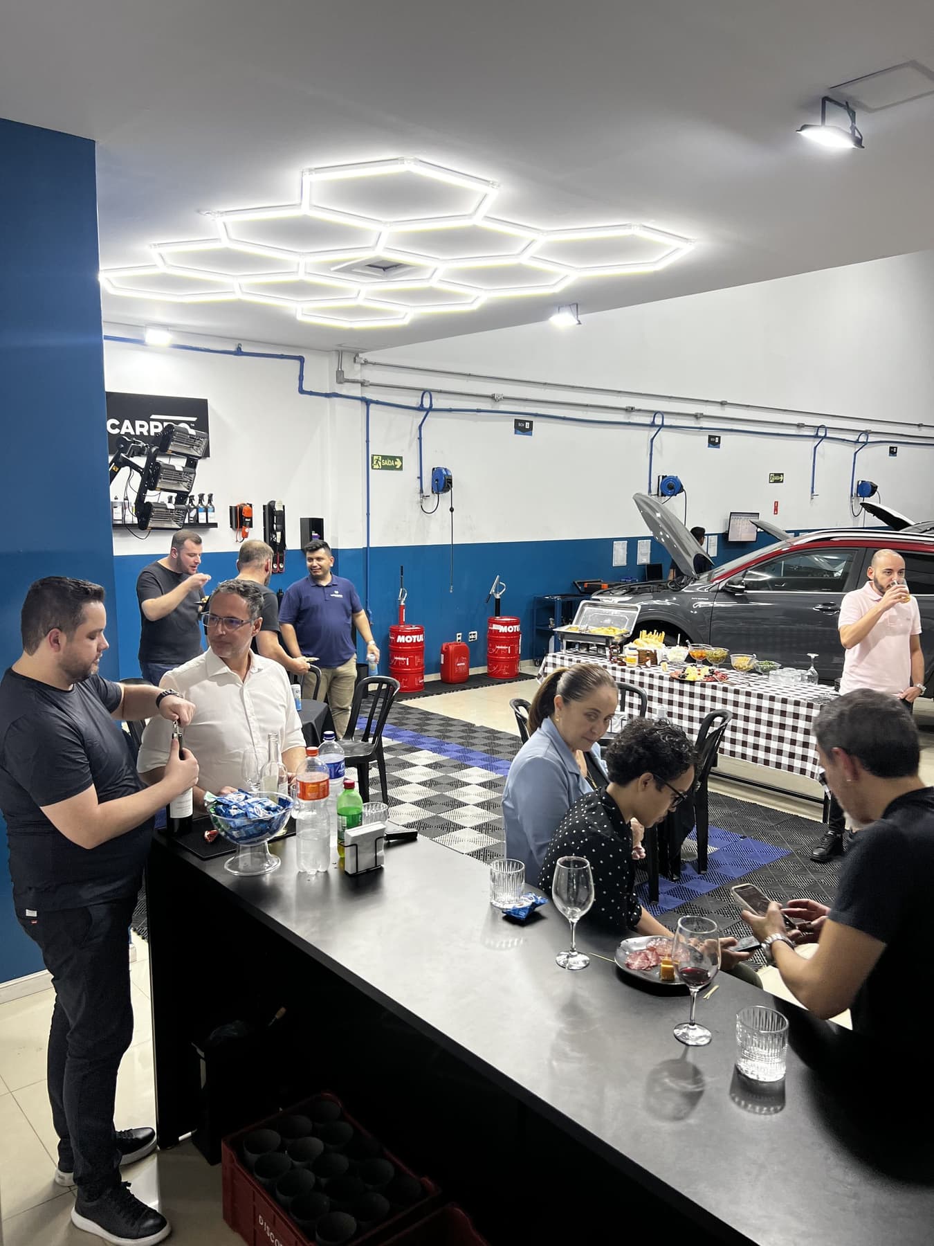 Convidados do UtaBar em volta do balcão montado no box da Utacar, com a iluminação hexagonal acesa e um carro em atendimento ao fundo.