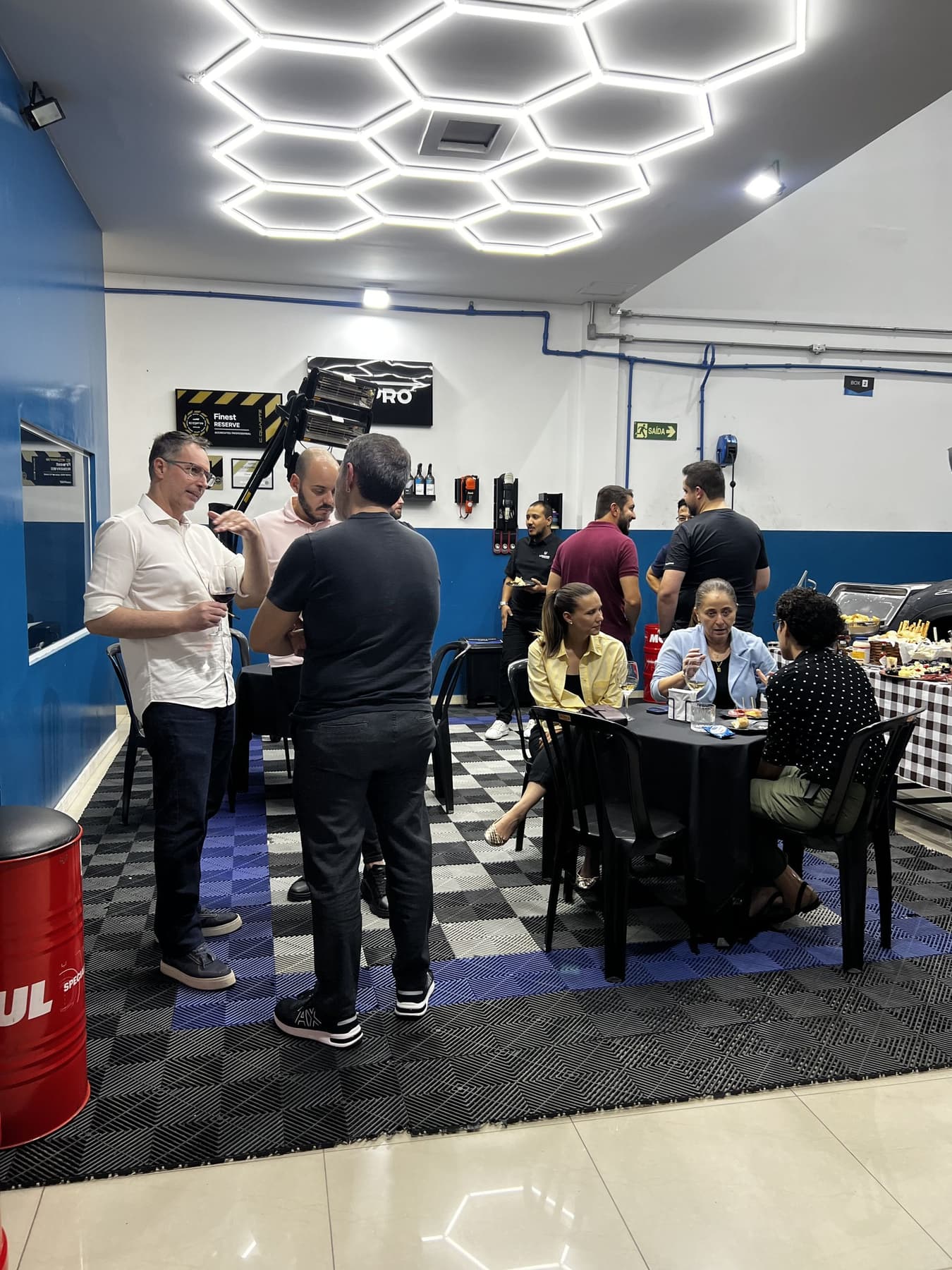 Mesas do UtaBar ocupadas por convidados no box da Utacar, com o buffet montado e a iluminação hexagonal acesa.