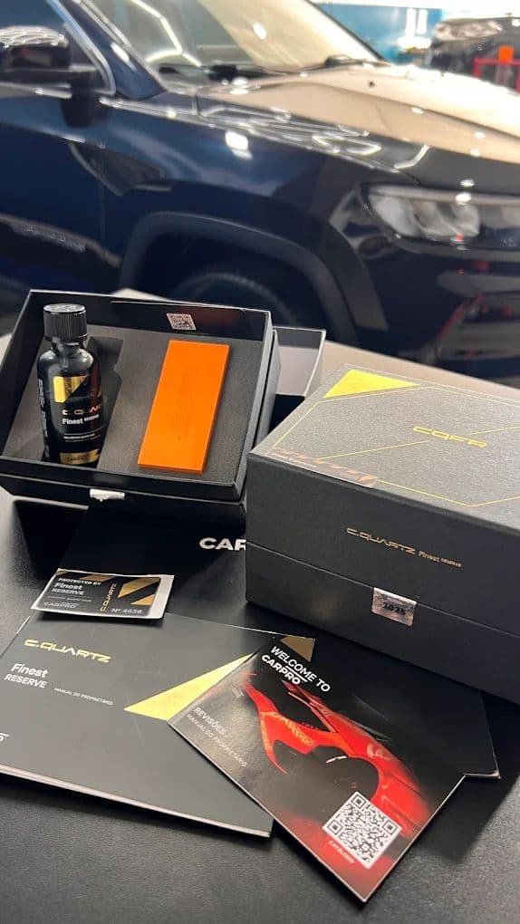 Kit CARPRO C.Quartz Finest Reserve aberto sobre a bancada, com o frasco, o aplicador e os manuais, e um Jeep escuro ao fundo.